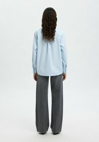 Camicia azzurra a maniche lunghe con taglio dritto, dotata di una spalla posteriore e polsini con bottoni, abbinata a pantaloni grigi a vita alta e gamba larga.