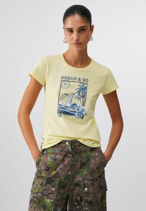 Mujer de cabello oscuro con una camiseta amarilla "Dream & Go" con un diseño de barco tropical, usando pantalones cargo de camuflaje verde con las manos en los bolsillos.