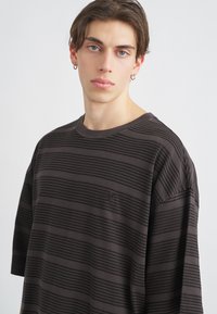 Camiseta de manga corta de color marrón oscuro con rayas, que presenta una textura acanalada, un cuello redondo y un pequeño detalle de logo en el pecho.