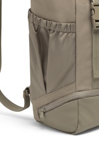 Khakinvärinen reppu, jossa on teksturoitu kangas, sivulla verkkotasku ja vetoketjullinen alaosaston lisäsäilytystä varten. Säädettävä olkahihna.