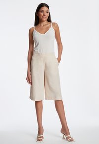 Culotte-Shorts aus hellbeigem Stoff, mit weitem Bein, seitlichen Taschen und einer glatten Textur. Kombiniert mit einem taillierten weißen Oberteil.