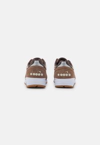 Ett par bruna mockaskor med vita sulor, sett från baksidan, med en vit logotyp på varje häl mot en enfärgad ljus bakgrund.