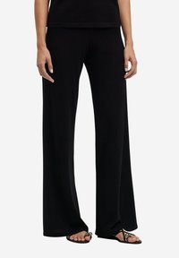 Zwarte wijde broek van zacht materiaal, met een relaxte pasvorm en afgewerkte zoom. Gecombineerd met eenvoudige zwarte sandalen.
