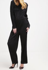 Mono negro con pliegues, de mangas largas, cintura ajustada y piernas anchas, confeccionado en una tela texturizada. Combinado con sandalias de tacón alto negras.