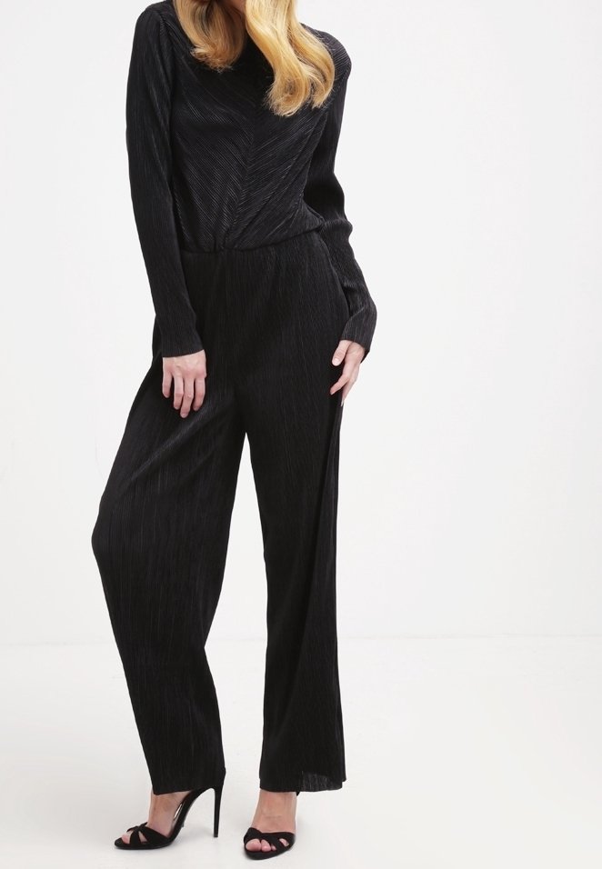 Mono negro con pliegues, de mangas largas, cintura ajustada y piernas anchas, confeccionado en una tela texturizada. Combinado con sandalias de tacón alto negras.