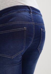 Jeans de denim azul oscuro con un diseño ajustado, que muestran dos bolsillos traseros y una costura en contraste a lo largo de las costuras.