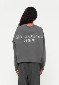 Grijze sweatshirt met ronde hals, drop shoulders, en witte "Marc O'Polo DENIM" op de achterkant gedrukt. Draagt een grijze geblokte broek.