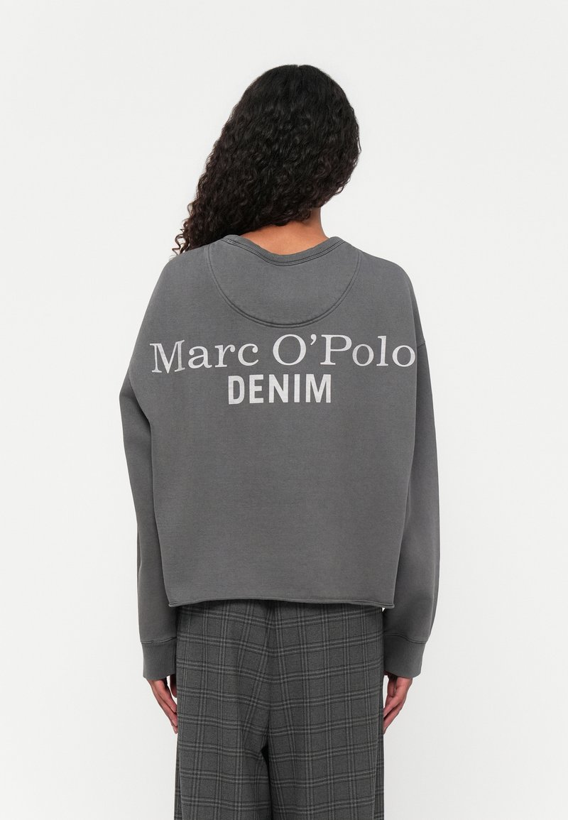 Grijze sweatshirt met ronde hals, drop shoulders, en witte "Marc O'Polo DENIM" op de achterkant gedrukt. Draagt een grijze geblokte broek.