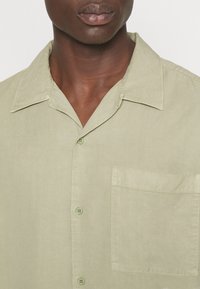 Chemise à boutons vert clair avec col, poche avant et tissu doux au toucher. Ourlet arrondi et détails de couture subtils visibles.
