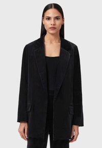 Una donna con capelli scuri e lisci indossa un blazer di velluto nero sopra un top a coste nero, in piedi di fronte a uno sfondo chiaro e uniforme.