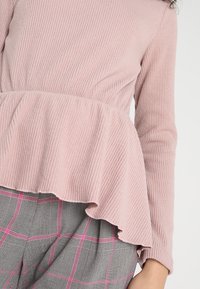 Rosa geripptes Oberteil mit langen Ärmeln und einem Peplum-Saum, kombiniert mit grauen karierten Hosen mit rosa Akzenten und einer strukturierten Oberfläche.