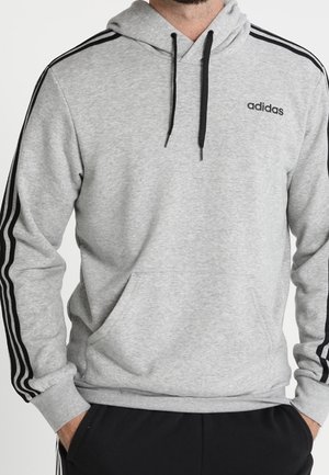 Sudadera gris de tejido suave con un bolsillo canguro, cordones negros y tres bandas negras que recorren las mangas. Logotipo "adidas" en el pecho.