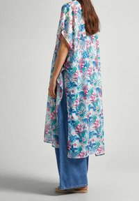 Kvinna iklädd en blommönstrad, ljusblå kimono över vida blå jeans och sandaler, stående på sidan mot en neutral bakgrund.