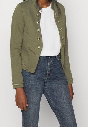 Personne portant une veste vert olive avec des boutons dorés sur une chemise blanche et un jean bleu, montrée des épaules jusqu'aux hanches.