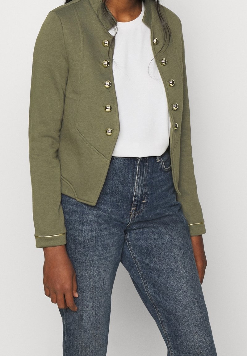 Personne portant une veste vert olive avec des boutons dorés sur une chemise blanche et un jean bleu, montrée des épaules jusqu'aux hanches.