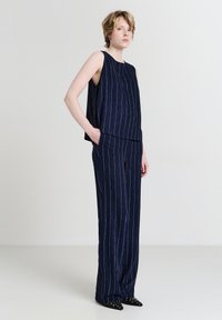 Femme debout les mains dans les poches, portant un haut sans manches marine à rayures et un pantalon large assorti avec des chaussures noires à talons pointus.
