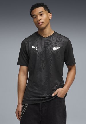Jeune homme portant un t-shirt de sport noir Puma avec un motif discret de fougère et des logos blancs, debout avec une main dans la poche sur un fond gris.