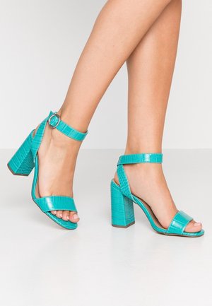 Jambes portant des sandales à talons blocs turquoise avec des brides de cheville et une texture crocodile sur un sol blanc lisse.