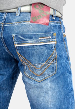 Jeans en denim bleu avec une finition texturée, des coutures orange et grises contrastées, et une étiquette de marque rouge et verte sur la taille.