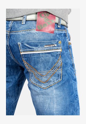 Jeans en denim bleu avec une finition texturée, des coutures orange et grises contrastées, et une étiquette de marque rouge et verte sur la taille.