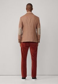 Brauner Tweed-Blazer mit Ellenbogenpatches, kombiniert mit rostfarbenen Cordhosen. Der Blazer hat einen Reverskragen und einen einzelnen Schlitz hinten.