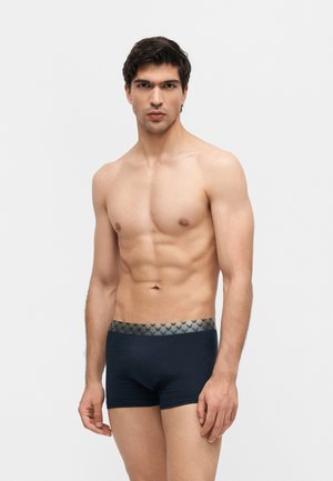 Noor lihaseline mees, kellel on tumedad bokserid mustrilise vöökohaga, seisab ühevärvilise heledas hallis taustal.