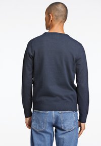 Pull en maille bleu marine avec un col rond et des poignets côtelés, porté sur un jean en denim bleu clair. La vue de dos montre un design d'épaule sans couture.