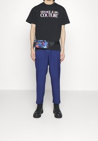 Osoba oblečená v černém tričku Versace Jeans Couture, modrých kalhotách, černých bojových botách a barevné ledvinkové tašce s černým popruhem.