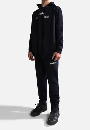 Sudadera con cremallera azul marino y joggers a juego, hechos de una tela suave, con estampados gráficos blancos en el pecho y en los pantalones; combinados con zapatillas negras.