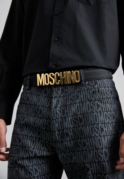 Černá košile s detaily na knoflíky, spárovaná s tmavými kalhotami s opakujícím se vzorem loga. Zlatý pásek "MOSCHINO" je výrazně viditelný.