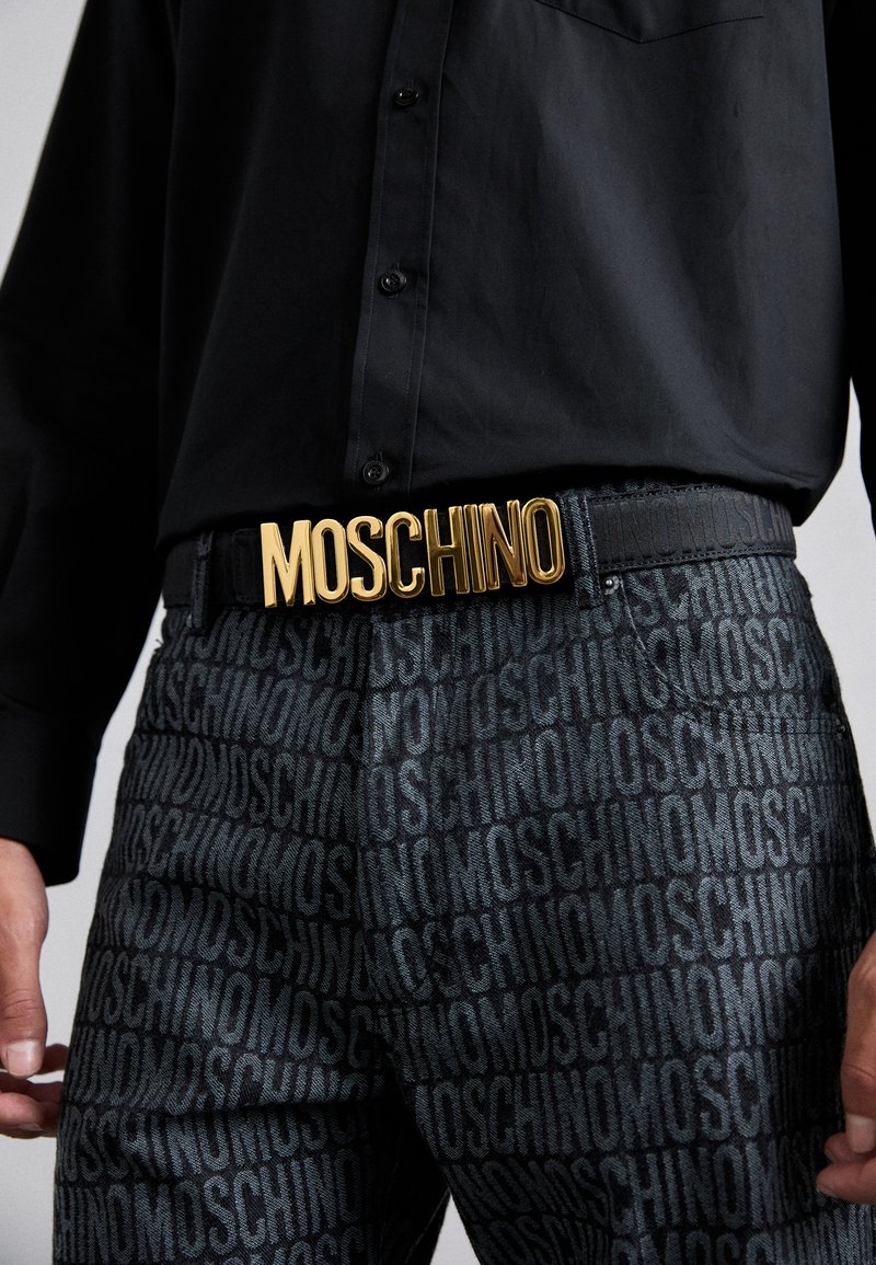 Črna majica z detajli gumbov, v kombinaciji s temnimi hlačami z ponavljajočim se logotipom. Zlati pas "MOSCHINO" je vidno izpostavljen.
