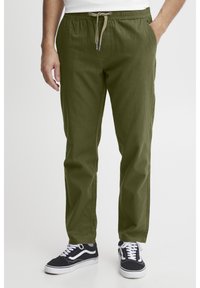 Mies, jolla oli päällään oliivinvihreät nyörivahteiset housut ja mustavalkoiset lenkkarit, toinen käsi taskussa, seisoo valkoista taustaa vasten.