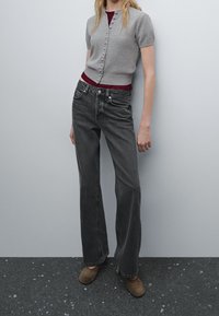 Cardigan grigio a maniche corte con dettagli a bottoni, indossato sopra una maglietta bordeaux. Jeans neri a gamba larga e stivaletti con tacco marroni completano l'outfit.