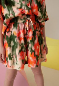 Copil îmbrăcat cu pantaloni scurți lejeri, tie-dye, și un top asortat în culori portocalii, verzi și crem, stând în fața unui fundal pastel.