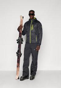 Melna slēpošanas jaka ar neona zaļiem akcentiem, melnas slēpošanas bikses, turētu koka slēpes ar stiprinājumiem un valkājošiem tumšiem saulesbrillēm.