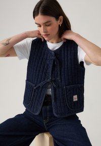 Kuidutud tumesinine denimvest sidekinnitusega, ribakujuline tekstuur ja kaks eesmist taskut; esitleb brändi märgist paremas taskus.