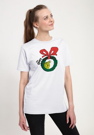 T-shirt bianca in cotone con una grafica del Grinch all'interno di una corona verde con dettagli rossi e un grande fiocco rosso in cima.