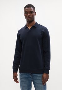 Tengerészeti kék, hosszú ujjú pólóing puha anyagból, galléros nyakkal és bordás mandzsettákkal. Világoskék farmernadrággal párosítva.