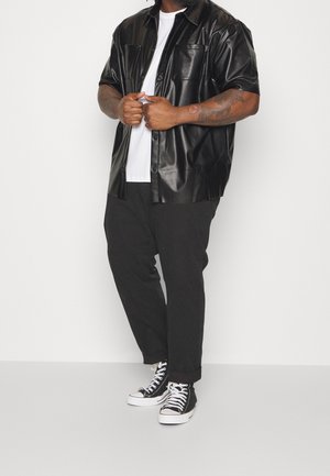 Schwarzes kurzärmeliges Hemd aus Kunstleder mit durchgehender Knopfleiste und zwei Brusttaschen. Kombiniert mit schwarzen Hosen und schwarzen High-Top-Sneakern.
