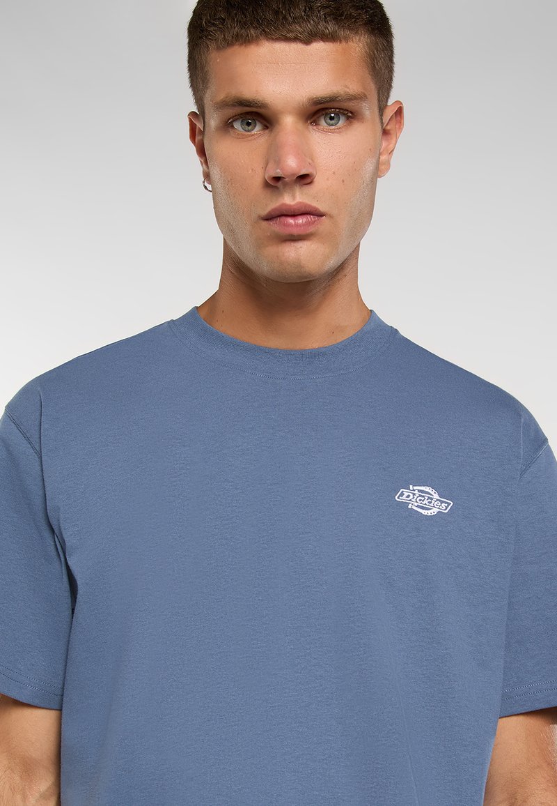 T-shirt di cotone blu con scollatura a girocollo, con un piccolo logo bianco sul petto. Il tessuto ha una consistenza morbida.