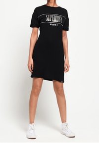 Robe t-shirt noire à manches courtes avec le logo "SUPERDRY" en blanc et argenté sur la poitrine ; coupe droite ; portée avec des baskets blanches.