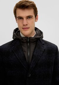 Donker geruite overjas met een afneembare zwarte gewatteerde hoodie. Voorzien van een rits en knoopsluiting aan de voorkant, en een textuurafwerking van de stof.