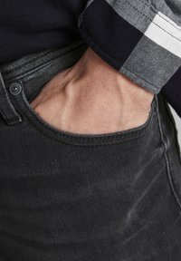 Mano metida en el bolsillo delantero de unos pantalones vaqueros negros, vistiendo una camisa de manga larga a cuadros negro y blanco.