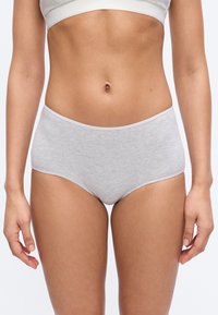 Γυναίκα που φοράει ανοιχτόγριζα βαμβακερά σλιπ ψηλής μέσης και ένα λευκό κοντό τοπ, στέκεται μπροστά σε απλό φόντο.