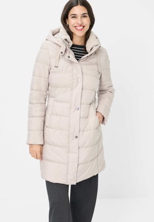 Smiling woman wearing beige quilted hooded coat with snap buttons, hand in pocket, and dark pants against white background.