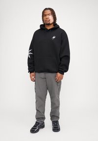 Felpa nera con cappuccio e tasca frontale, decorata con un logo a ragno, abbinata a pantaloni cargo grigi. Le calzature presentano una base nera con accenti viola.