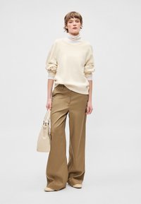 Sudadera color crema con mangas abullonadas, combinada con pantalones anchos de color caqui. Se sostiene un bolso beige en la mano derecha. Texturas suaves en todo el conjunto.