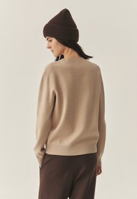 Maglione beige lavorato a maglia con orlo a coste, colletto rotondo e maniche lunghe, abbinato a pantaloni marroni e un berretto a coste marrone.