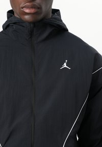 Veste à capuche noire en matériau léger, dotée d'une fermeture éclair et d'un passepoil blanc contrastant. Comprend un logo sur la poitrine.