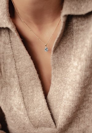 Goldene Halskette mit einem blauen, birnenförmigen Anhänger, getragen über einem hellbeigen, gestrickten Pullover mit tiefem V-Ausschnitt.